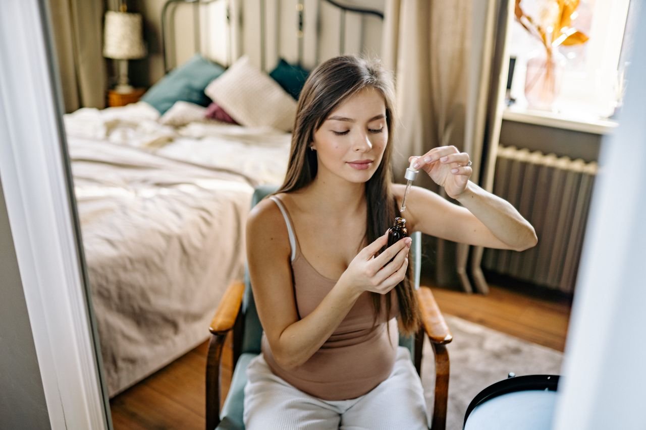 About A woman in a tank top applies skincare serum while seated in a comfortable bedroom.