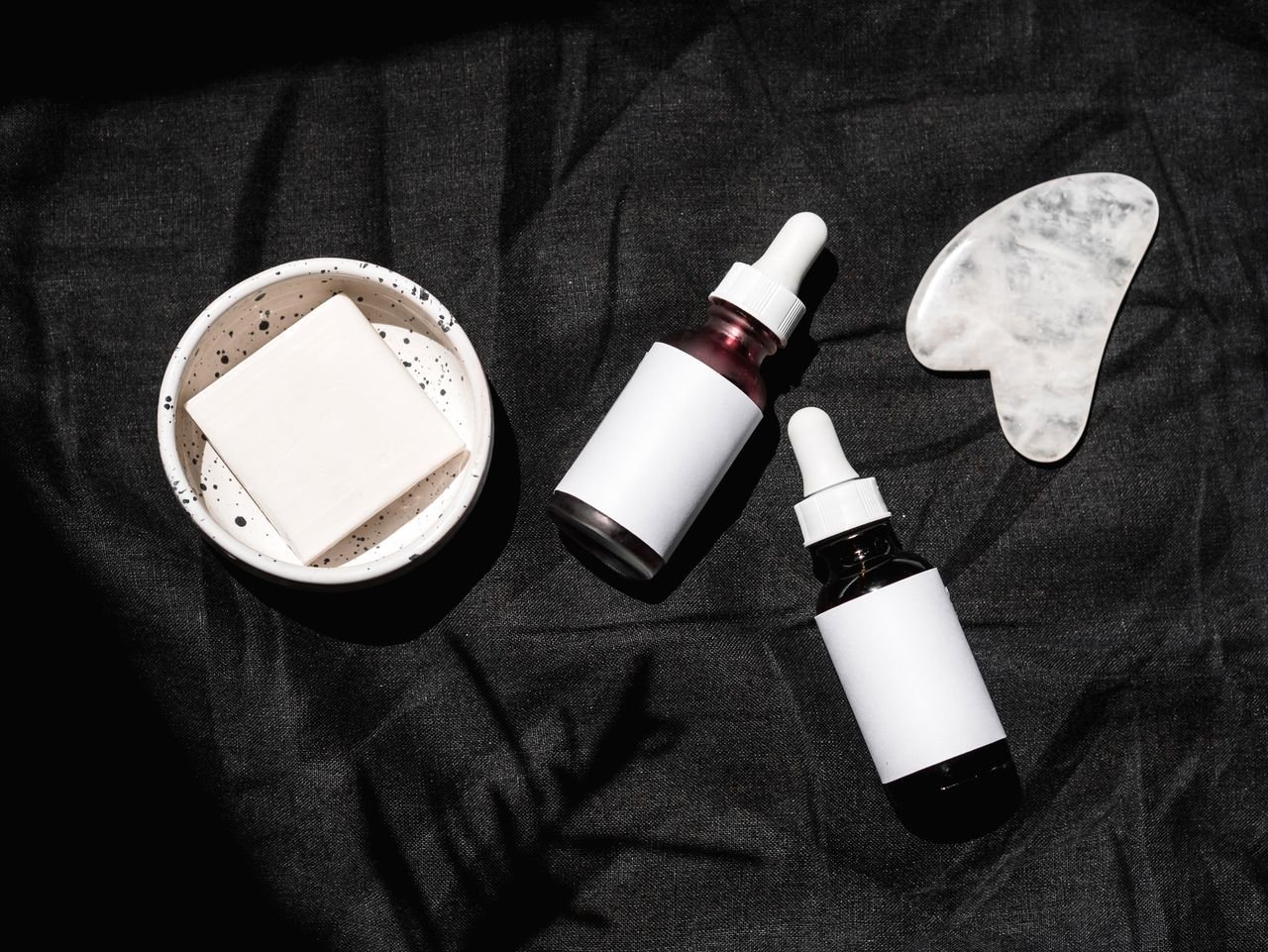 High-angle view of skincare products including serums, gua sha, and bar soap on a dark fabric backdrop.